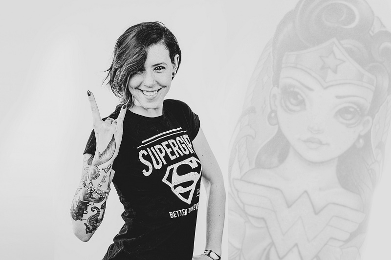 Getatoeëerde vrouw met Supergirl-shirt maakt rockgebaar; naast haar een grote, vervaagde Wonder Woman-illustratie, zwart-wit.