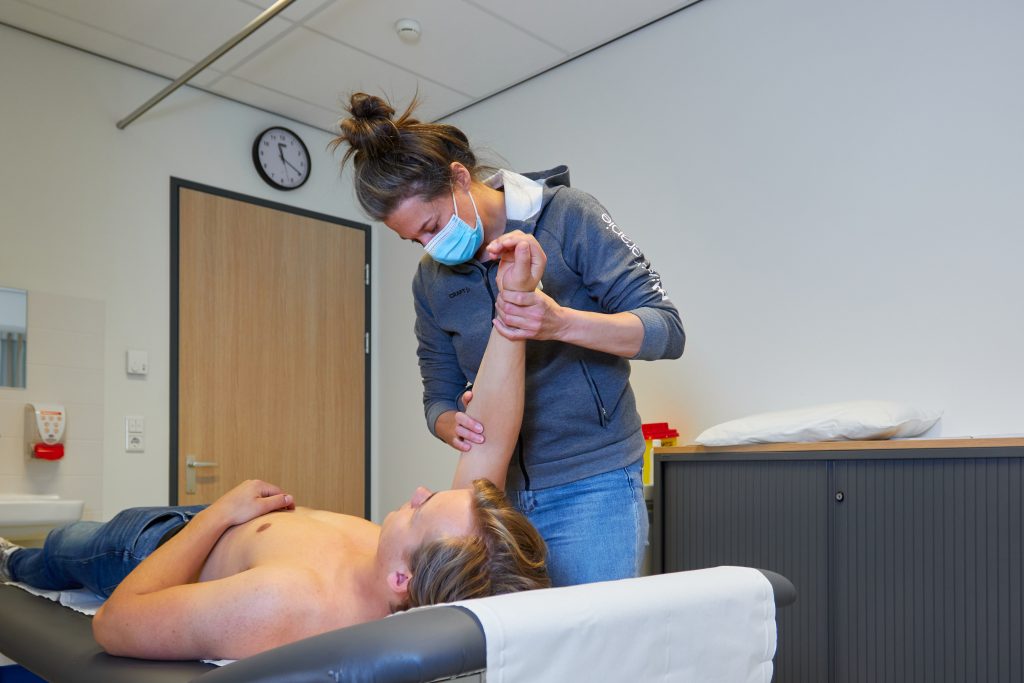 Een gemaskerde persoon voert fysiotherapie uit en tilt de arm van een shirtloze patiënt op in een klinische ruimte.