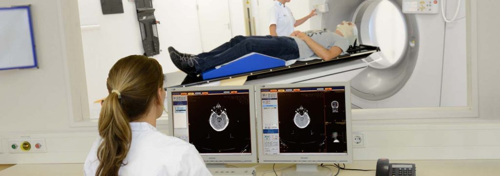 Een patiënt ligt op een CT-scanner. Een technicus bereidt hoofdbehandeling voor; een ander bewaakt scans op computerschermen.