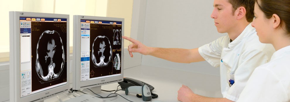 Twee artsen in witte jassen bekijken en bespreken CT-scans van longkanker en radiotherapie op twee monitoren; één wijst iets aan.