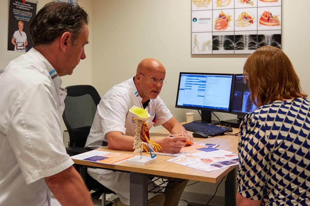Drie zorgprofessionals bespreken thoracic outlet syndroom bij een bureau met medische dossiers, modellen en computerschermen.