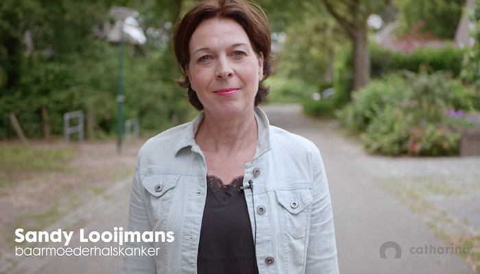 Vrouw met kort bruin haar en spijkerjack op bospad. Tekst: Sandy Looijmans, symptomen en vooruitzichten baarmoederhalskanker. Catharina logo rechtsonder.