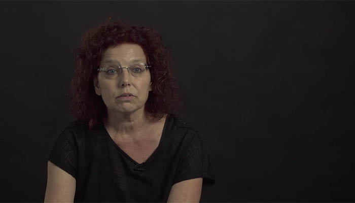Persoon met krullend rood haar, bril en zwart shirt zit voor donkere achtergrond en kijkt neutraal in de camera; thema endeldarmkanker.