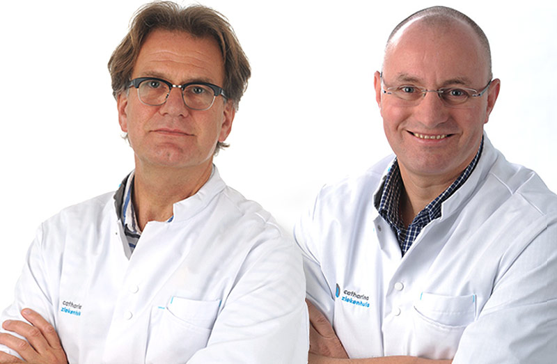 Twee mannen in witte labjassen en brillen, de een met bruin haar en serieus, de ander kaal en glimlachend, staan naast elkaar.
