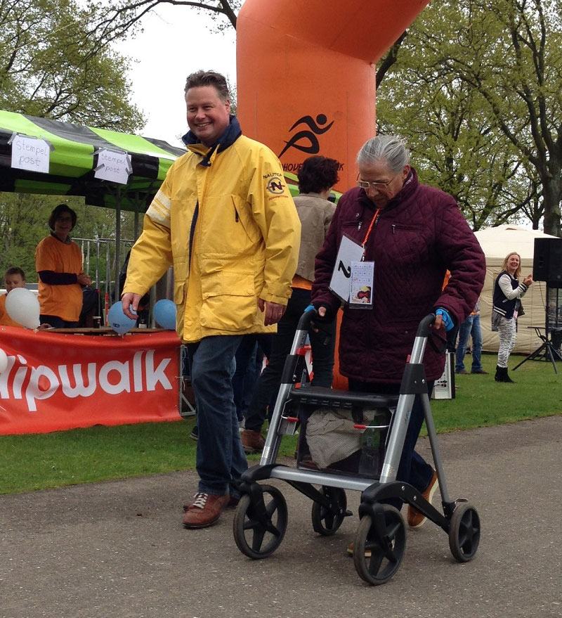 Man in gele jas loopt naast oudere vrouw met rollator bij HipWalk Nederland, onder oranje boog met mensen en rode banner.