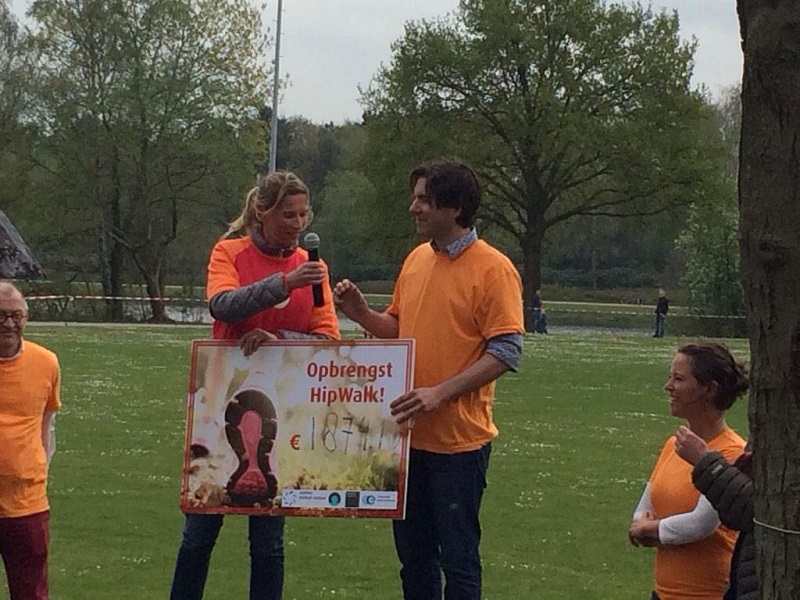 Twee mensen in oranje shirts tonen een cheque van €1874 voor HipWalk Nederland, buiten op gras met anderen en bomen achter hen.