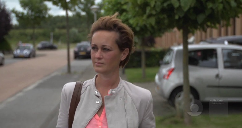 Een persoon met kort bruin haar en lichte jas loopt met een bruine tas langs geparkeerde auto's en bomen op een bewolkte dag.