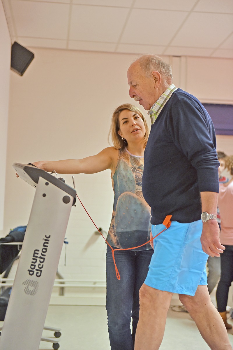Vrouw begeleidt oudere man met etalagebenen op daum electronic-loopband in fitness- of revalidatiecentrum.