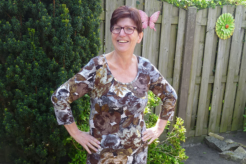 Vrouw met kort bruin haar en bril staat buiten bij schutting met groen, glimlacht en heeft handen op heupen. Roze vlinder op schutting.