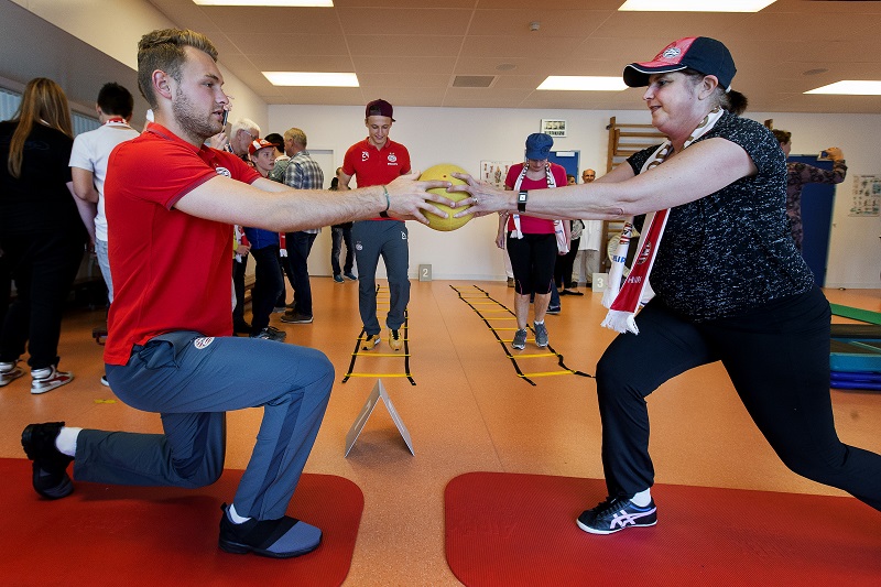 Man en vrouw balanceren op één knie op matten en houden samen een gele bal vast in een sportschool voor borstkankerpatiënten.