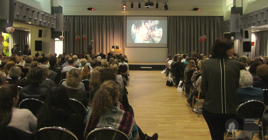 Een groep mensen, waaronder hartpatiënten, kijkt naar een presentatie van Paul Engels in een conferentiezaal met grijze gordijnen.