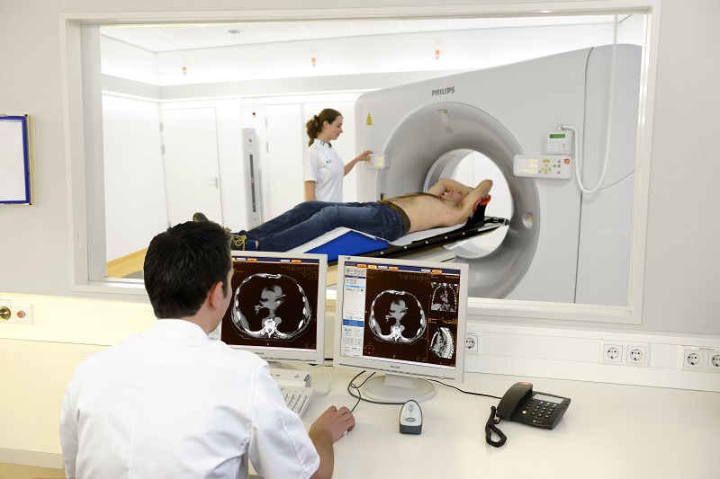 Een man ligt op een CT-scanner, een technicus bereidt het apparaat voor. In de controlekamer worden de scans gemonitord.