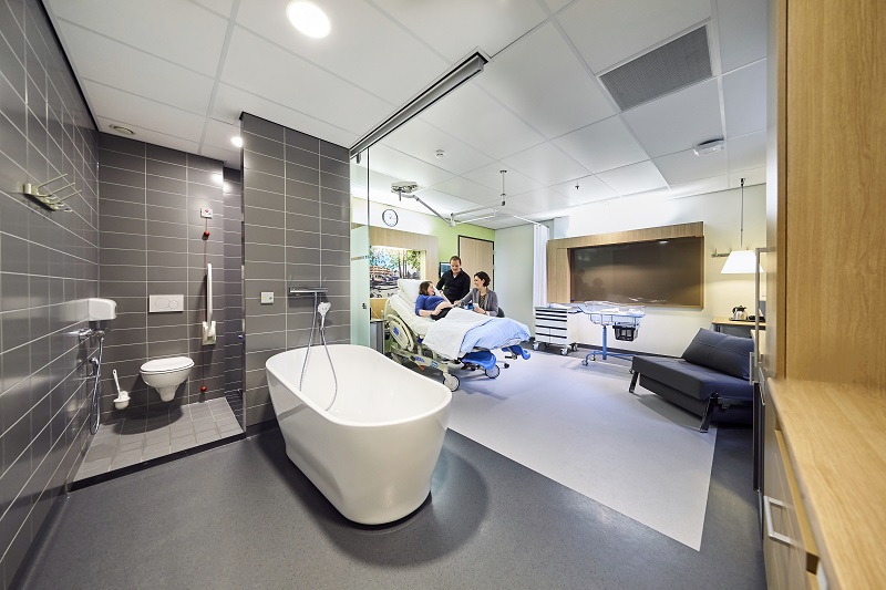 Moderne kraamsuite met bevalbad op de voorgrond, open badkamer links en medisch personeel bij patiënt op bed op de achtergrond.
