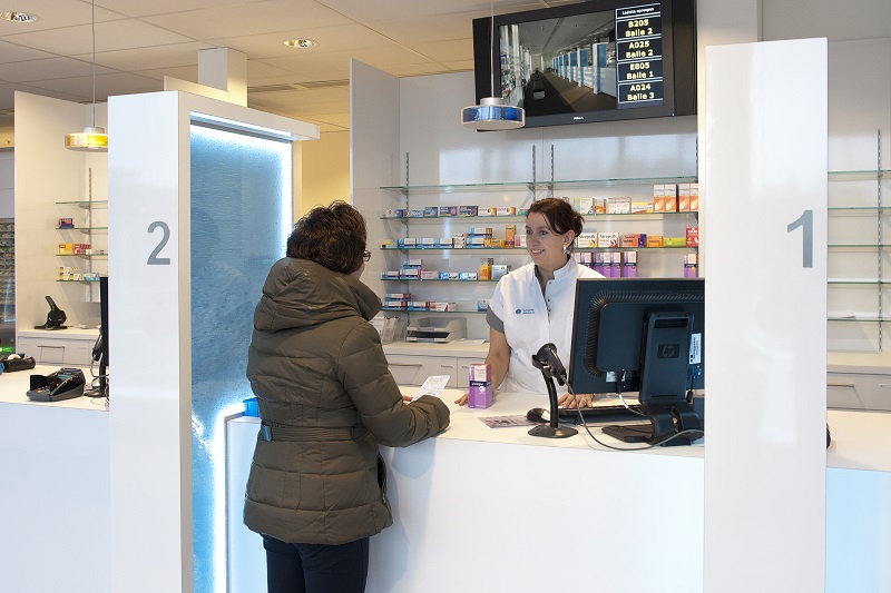 Vrouw in groene jas bij apotheekbalie spreekt met medewerker in witte jas; medicijnkasten en computerscherm op de achtergrond.