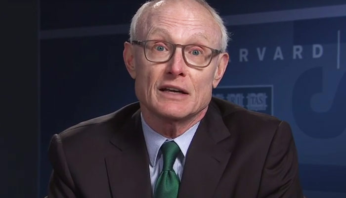 Man met lichte huid, bril en kort licht haar in donker pak met witte blouse en groene stropdas, voor blauwe HARVARD-achtergrond.
