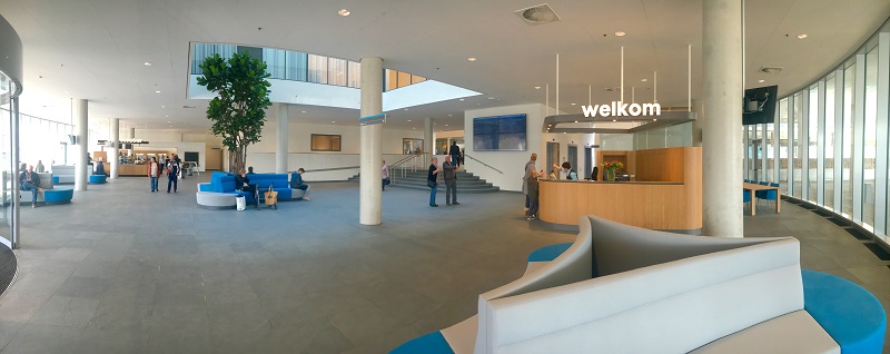 Ruime, moderne lobby met grote ramen, welkom-bord, balie, mensen, blauw-grijze zitplaatsen en grote binnenboom bij hoofdingang.