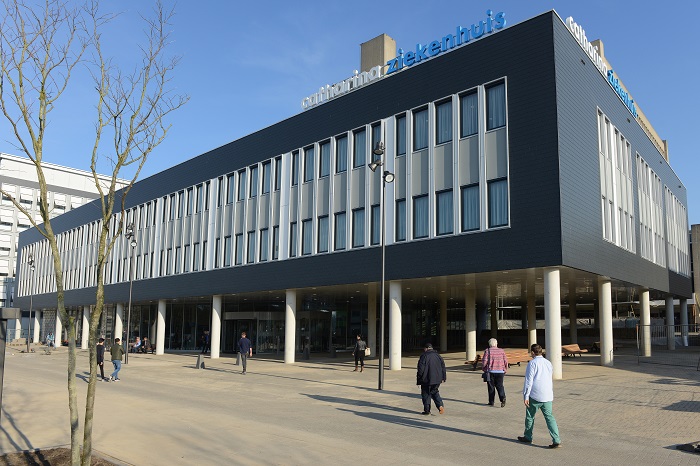 Modern ziekenhuis met grote ramen en bord 'ziekenhuis'. Mensen lopen buiten bij de ingang op een zonnige dag.