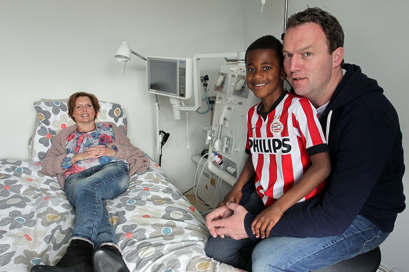 Janneke Bresser ligt glimlachend op een ziekenhuisbed. Een man zit ernaast met een jongen in marathonshirt op schoot.