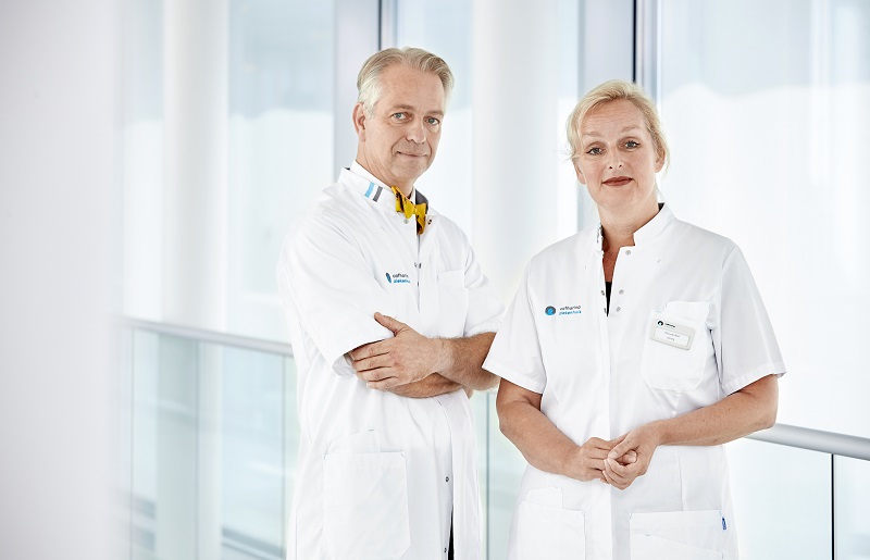 Twee artsen, man en vrouw, staan naast elkaar in een verlichte bende. Ze dragen witte jassen en kijken neutraal naar de camera.