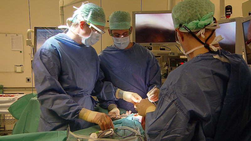 Drie artsen in OK-kleding voeren een operatie uit, omgeven door medische apparatuur en beeldschermen.