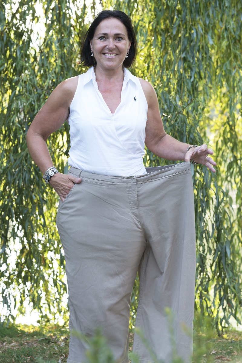 Vrouw buiten voor groen, glimlachend en met grote kaki broek; toont duidelijk haar succesvolle gewichtsverlies.