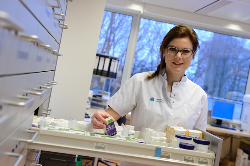 Vrouw in witte jas bij lade met medische spullen, houdt container vast en kijkt naar camera in modern laboratorium of ziekenhuis.