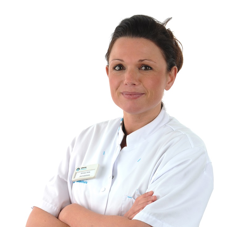 Vrouw in wit medisch uniform met gekruiste armen, donker haar en naamplaatje waarschuwt voor huidkanker. Witte achtergrond.