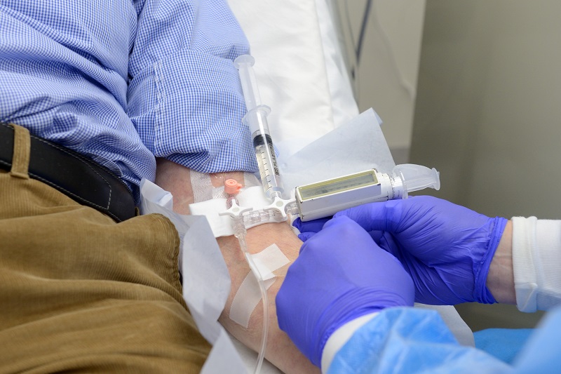 Een zorgverlener met paarse handschoenen dient medicatie toe via infuus in de hand van een patiënt met blauw geruit overhemd.