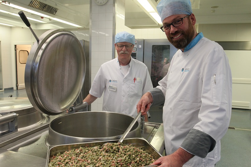 Twee mannen in witte jassen en blauwe haarnetjes staan in een keuken; één roert in een grote pan met eten.