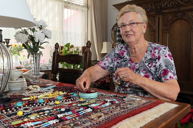 Een oudere vrouw zit aan een tafel met kleurrijke kralenkettingen. Ze glimlacht en pakt een sieraad op, omringd door bloemen.