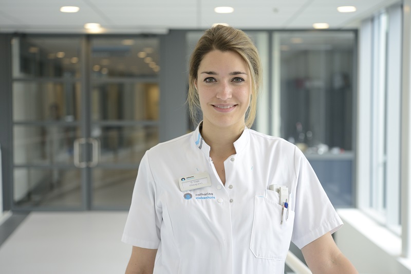 Vrouw in wit medisch uniform met badge en pennen in fel verlichte bende, klaar om borstkankerpatiënten te helpen.