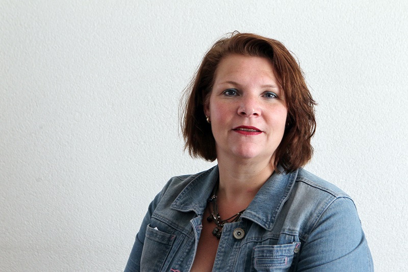 Vrouw met halflang bruin haar, spijkerjasje en ketting zit voor witte muur en kijkt met neutrale blik in de camera.