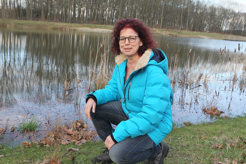 Persoon met krullend rood haar en bril hurkt bij vijver met riet en bomen op een bewolkte dag, draagt blauwe jas en jeans.