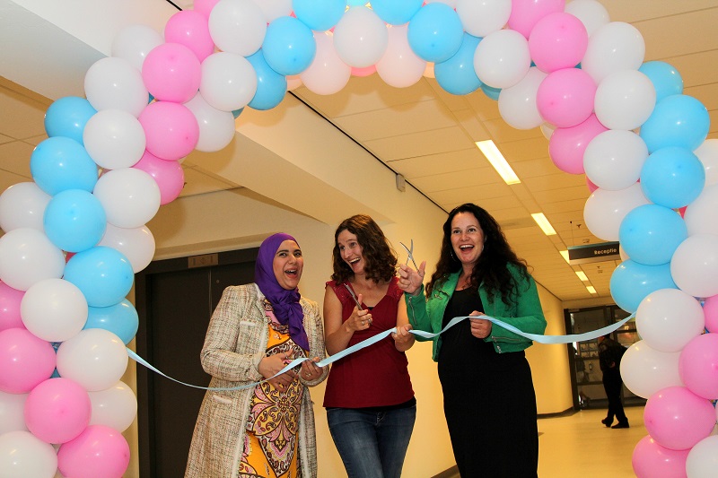 Drie vrouwen onder een roze, wit en blauwe ballonboog knippen samen een blauw pluisje bij de opening van nieuwe kraamsuites.
