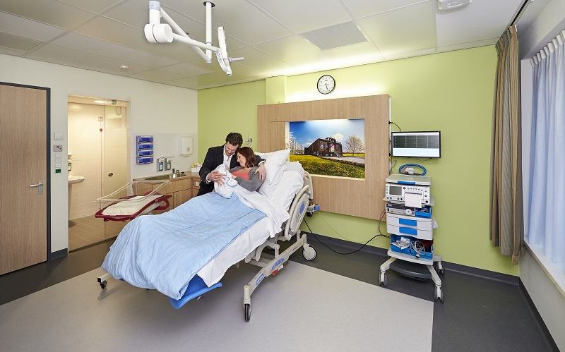 Vrouw met pasgeborene in bed, volwassene erbij. Medische apparatuur, klok en landschap aan de muur, badkamerdeur open.