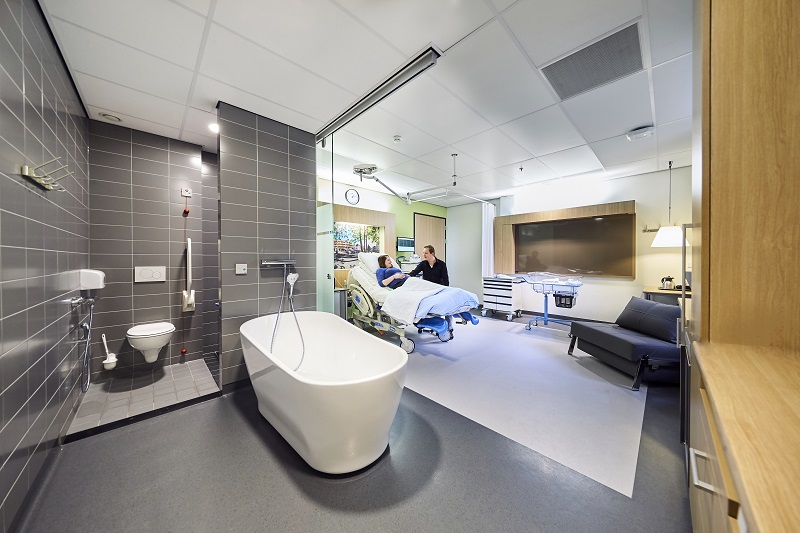Moderne kraamsuite met bad, rolstoeltoegankelijk toilet, bed voor bezoek, medische apparatuur en bank op de achtergrond.