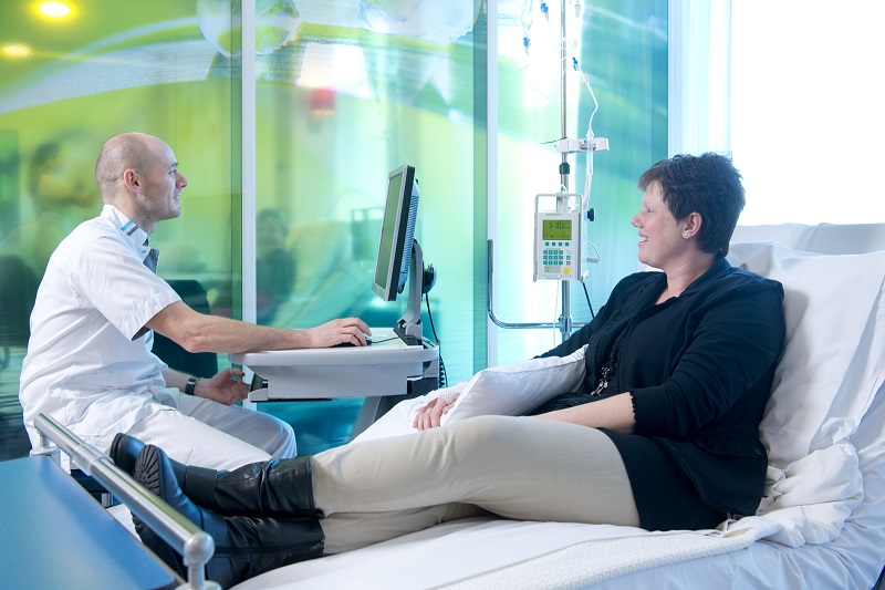 Een verpleegkundige bij een computer spreekt met een patiënt met infuus. Het team werkt samen voor betere zorg in de kliniek.