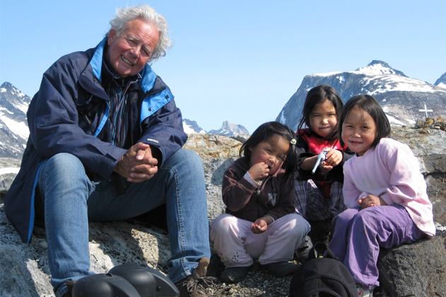 Een oudere man zit buiten op een rots met drie kinderen. Op de achtergrond besneeuwde bergen en een blauwe lucht.