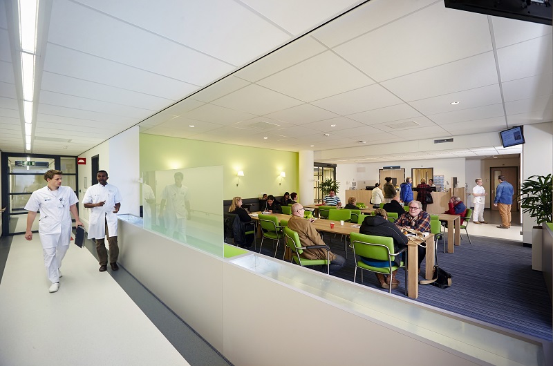 Moderne, lichte wachtkamer in ziekenhuis met mensen op groene stoelen. Twee medewerkers lopen door gang met planten en glas.