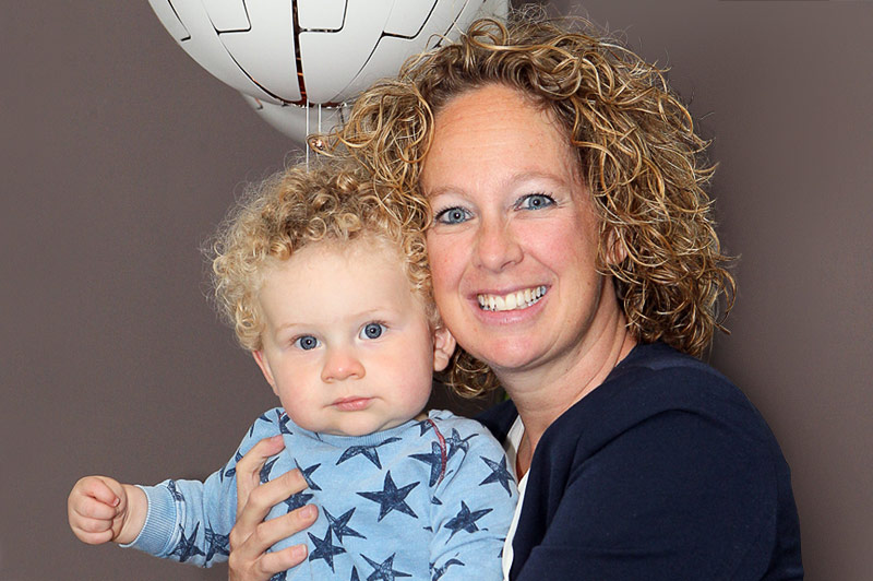 Vrouw met krullend haar houdt lachend een kind met blond krullend haar vast. Op de achtergrond een witte ballon en een muur.