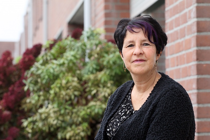 Vrouw met kort donker haar en paarse lok zit buiten bij een bakstenen gebouw, met zwart vest. Groene struiken en rode planten achter.