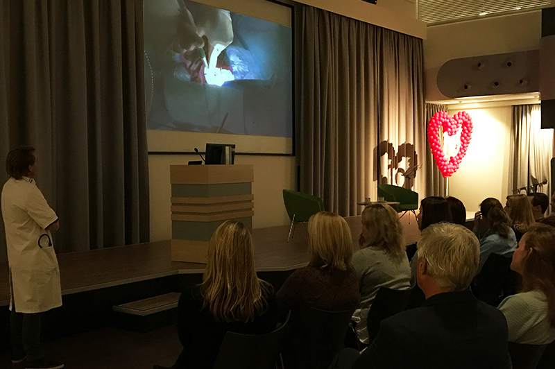 Groep kijkt in auditorium naar TAVI-behandeling op groot scherm; persoon in labjas en rode hartballonnen op de achtergrond.