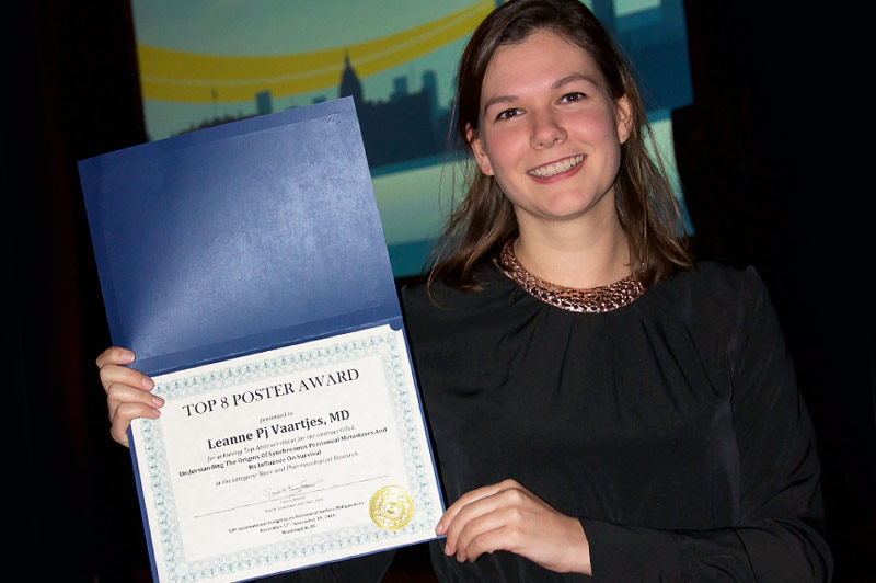 Op een congres toont iemand in zwart shirt een blauwe kaart met Top 8 Poster Award voor buikvlieskanker; scherm op achtergrond.