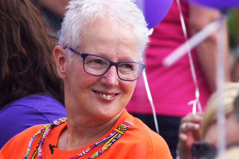 Oudere persoon met kort wit haar en bril glimlacht, draagt oranje shirt en kralenkettingen. Ballonnen en mensen op de achtergrond.