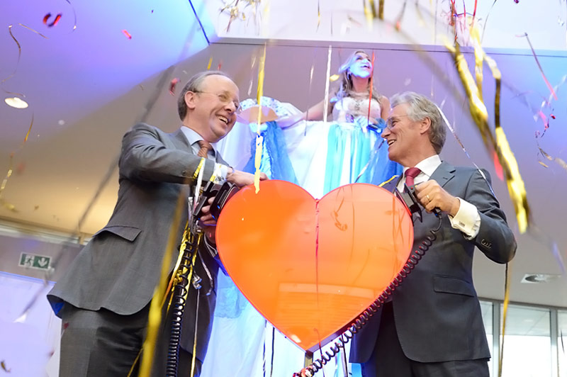 Twee mannen in pak houden kabels aan een groot rood hart vast. Confetti. Vrouw in witte jurk op podium bij Catharina Hartcentrum.