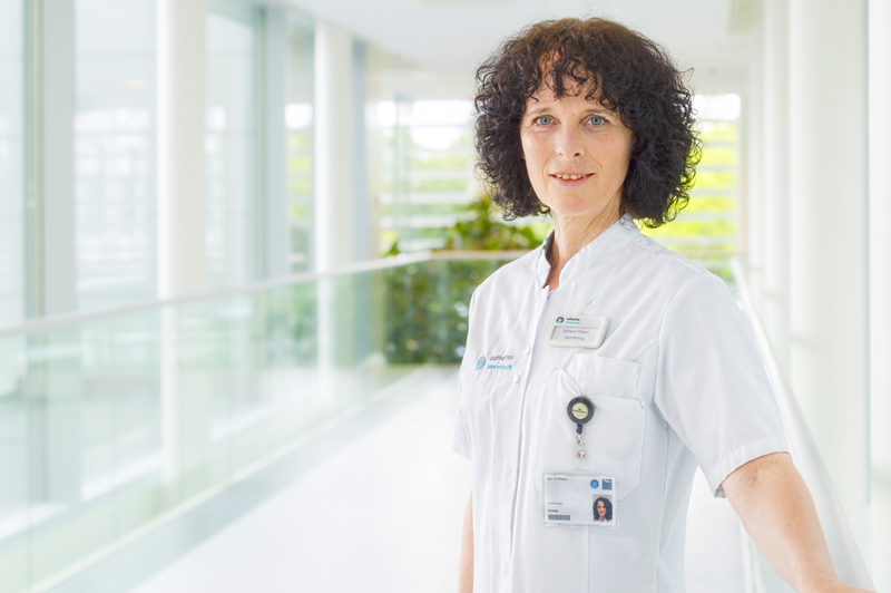 Vrouw met krullend donker haar in wit uniform en ID-badge staat glimlachend in lichte bende met glazen panelen.