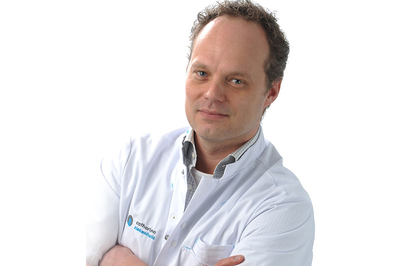 Man met kort, krullend haar in witte jas en geruit overhemd, armen over elkaar, kijkt naar camera tegen witte achtergrond.