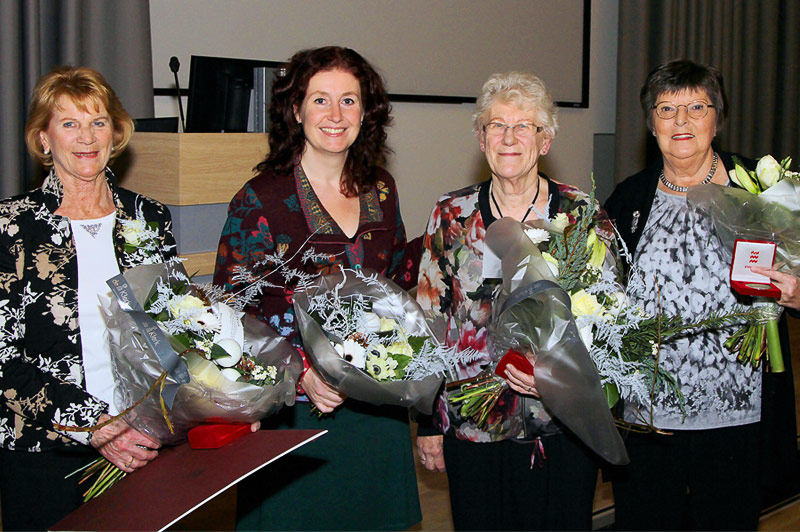 Vier vrouwelijke vrijwilligers poseren binnen met bloemen en cadeaus bij een podium en scherm, gekleed in nette kleding en lachend.