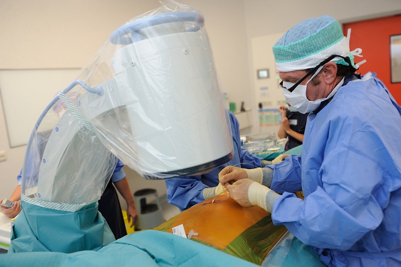 Een arts in OK-kleding voert met beeldapparatuur een procedure uit op een bedekte patiënt, met een assistent en instrumenten erbij.