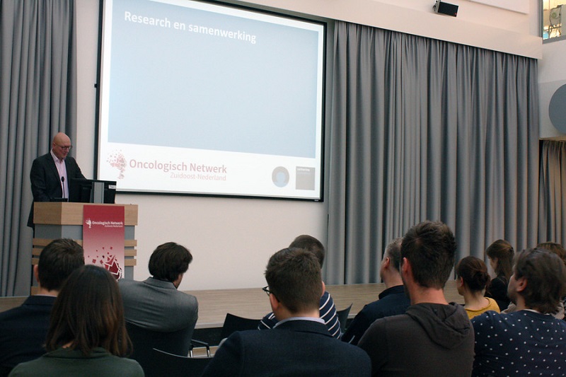 Een man geeft een presentatie bij een podium. Op het scherm staat "Research en samenwerking" en het logo van Oncologisch Netwerk Zuidoost Nederland.